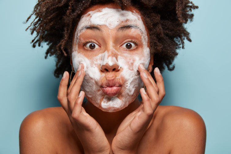People, wellness, hygiene and skin problems concept. Lovely Afro American lady keeps lips folded, touches cheeks, has white foam on faces, washes with beauty gel, feels refreshed, has wide opened eyes